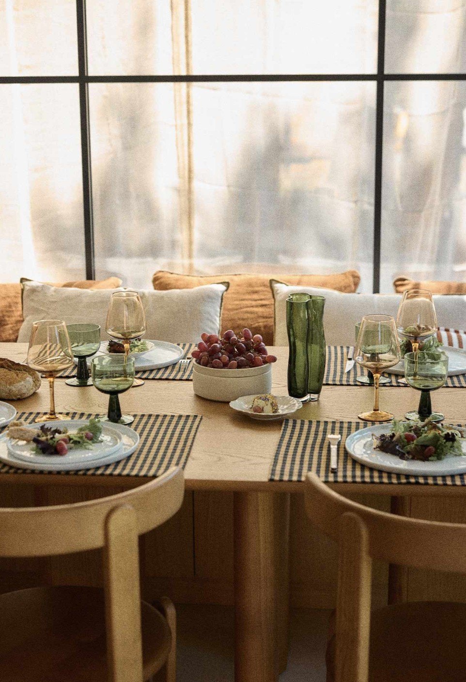 Table en bois dressée avec sets de table vichy, verres colorés et coussins en lin devant une verrière, élégance intemporelle de Margaret